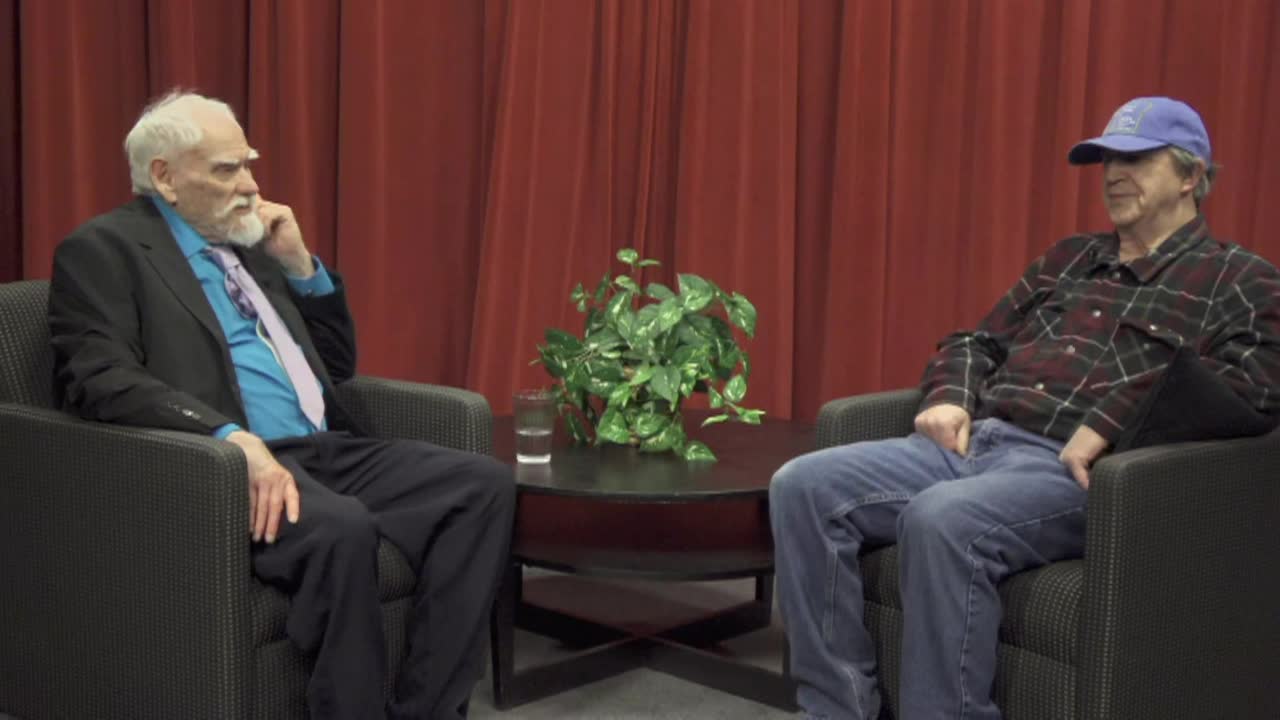 Red curtain background. Older man in a suit seated to the left and another older man seated to the right