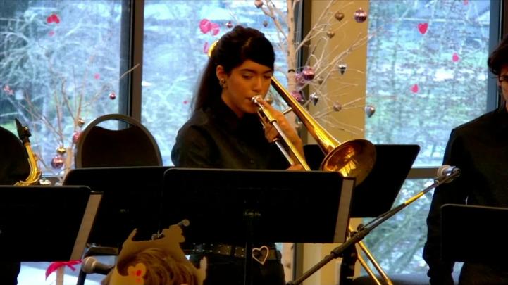 Student dressed in black standing in front of windows, playing a trombone 