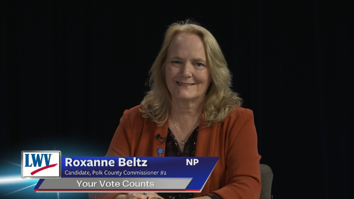 Black background. Woman with an orange suit jacket and black shirt seated in the middle. Blue and grey lower thirds and white words "Roxanne Beltz Candidate, Polk County Commissioner #2 Your Vote Counts"