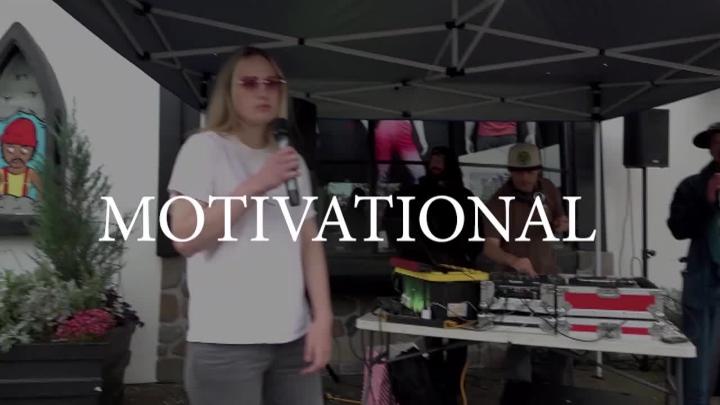 Blurred image of a woman in a white tee holding a mic. People standing in the background around a table with mixing equipment on it. Across the middle, white word "MOTIVATIONAL".