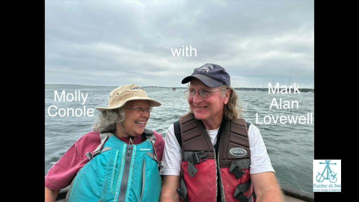 Older woman in a life jacket and hat seated on a boat with water in the background, to the left. Man in a brown life jacket, hat and glasses seated to the right.