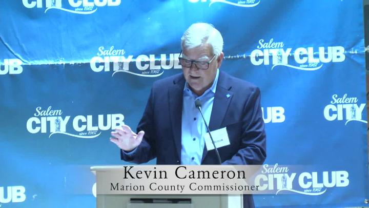Blue background with white Salem City Club words. Man wearing a blue suit speaking behind a podium. Lower thirds saying Kevin Cameron 