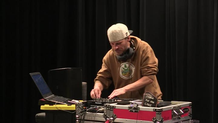 black background, dj in a tan sweatshirt and backwards hat on turntables