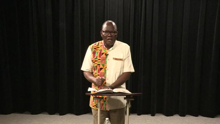 Black curtain background. African man in a cream and patterned shirt and glasses standing behind a small podium.
