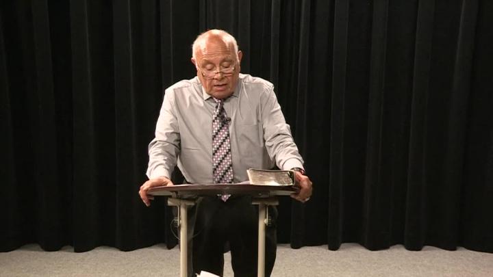 Black curtain background with an older man in a light colored button up and tie standing behind a smaller podium.