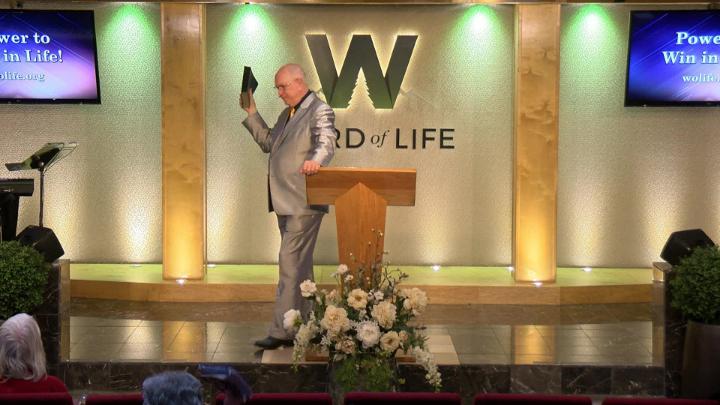 Man in a suit holding a bible next to a pulpit in the middle of a platform. Lit up white walls in the background with a large W and Word of Life in the center.