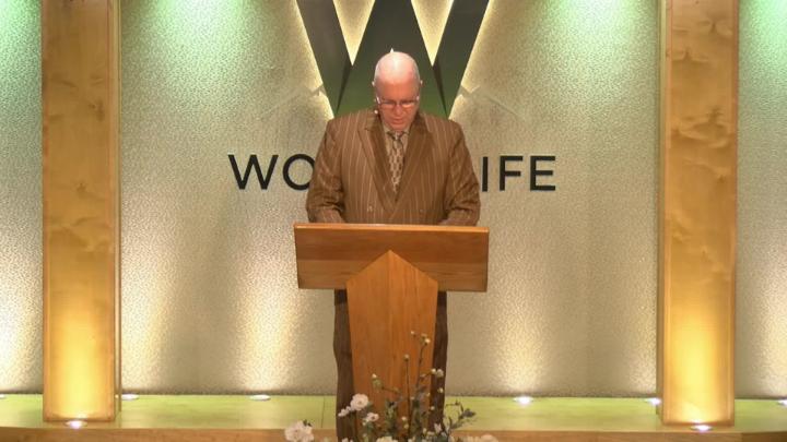 Man in a suit holding a bible next to a pulpit in the middle of a platform. Lit up white walls in the background with a large W and Word of Life in the center.