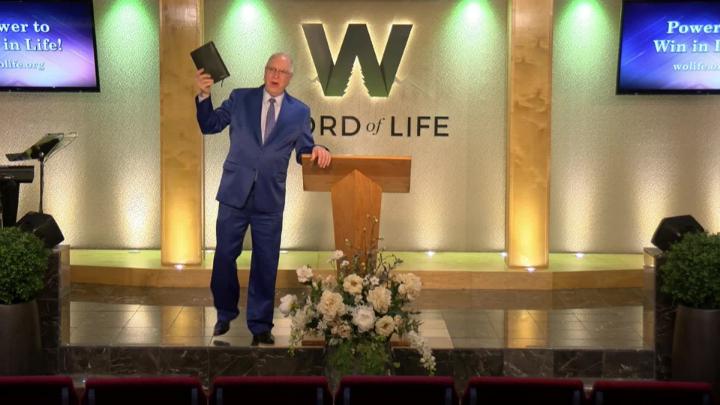 Man in a suit holding a bible next to a pulpit in the middle of a platform. Lit up white walls in the background with a large W and Word of Life in the center.