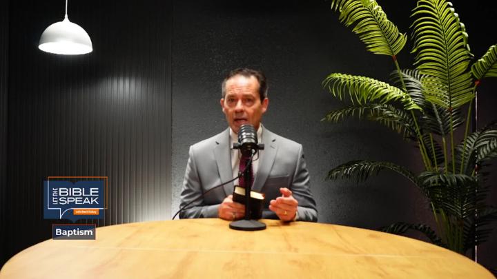 Man in a grey suit seated behind a table with podcast mic. Tall green plant to the back right. "Let the Bible Speak" logo bottom left.