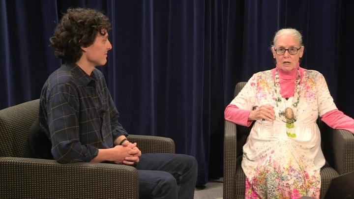 Dark curtained background. Older woman in a white and pink floral dress seated to the right. Man wearing dark clothes seated to the left.