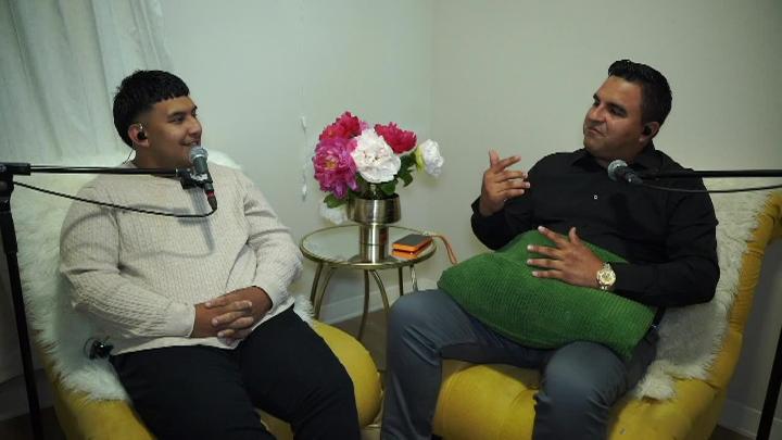 Two men seated in front of podcast mics. Man on left wearing white. Table with flowers in the middle. Man on right wearing dark clothing.
