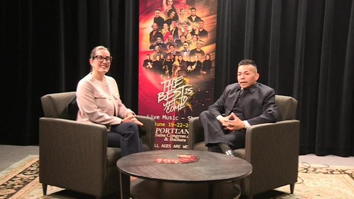 Black curtain background. Poster of dances in the back center. Man in dark clothing seated to the right. Woman in classes and tan jacket seated to the left.