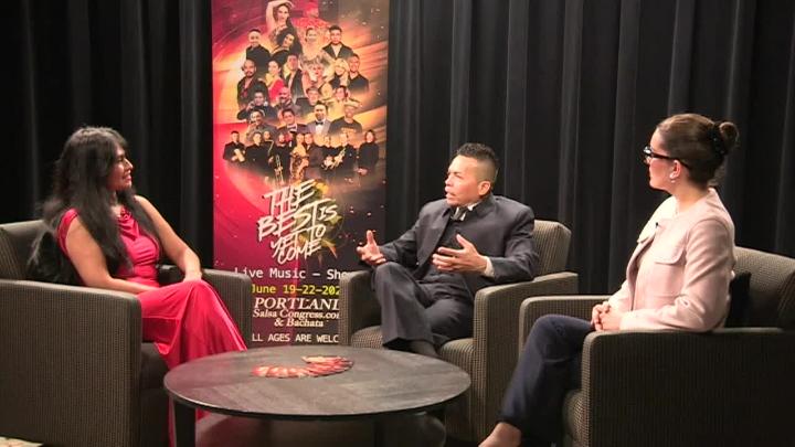 Black curtain background with a poster of salsa dancers on it. Woman in a red dress seated on the left. Man in dark clothing seated middle right and a woman in tan seated next to him