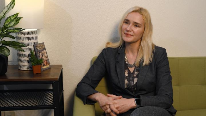 Blonde woman wearing dark clothing seated on a green couch. Table to the left with knick knacks on it.