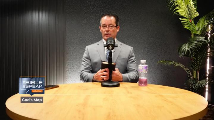 Man in a grey suit and glasses seated at a table behind a podcast mic. Plant to the right, Blue Let the Bible Speak logo bottom left.