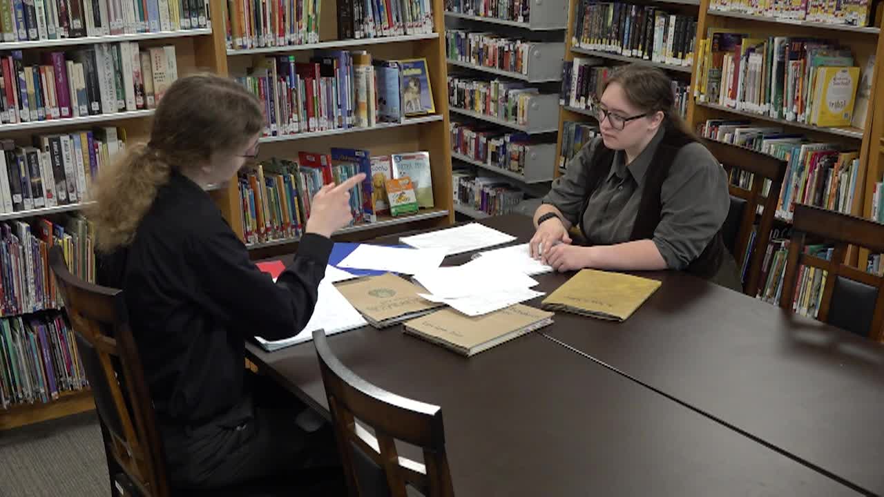 Two people in dark clothes seated at a table with lots of papers on it. Bookshelves surrounding them.