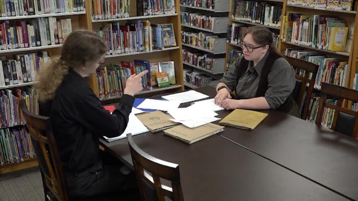 Two people in dark clothes seated at a table with lots of papers on it. Bookshelves surrounding them.