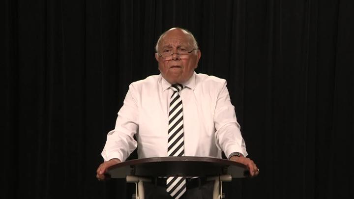 Black background. Older man in a button up shirt and tie standing in front of a podium.