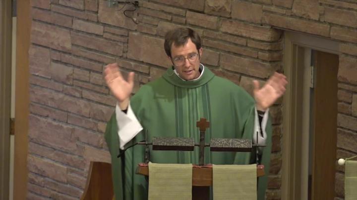 Priest dressed in green at a pulpit. Brick wall behind