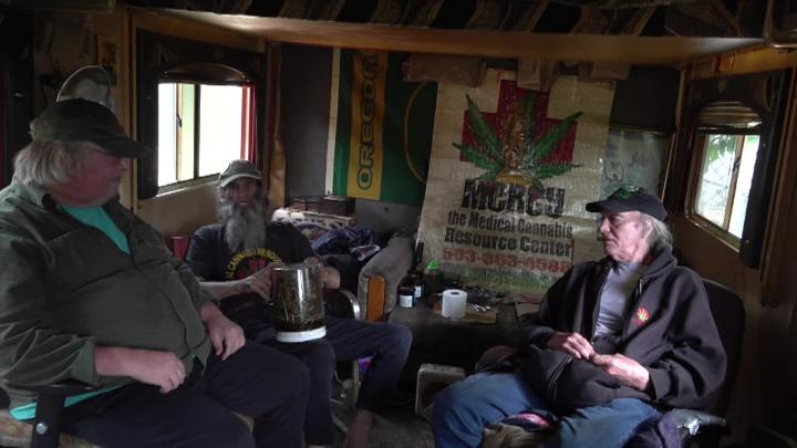 Man seated to the left in a green button up. Bearded man sitting in the back. Man in a jacket and hat seated to the right. Mercy logo sign in the background