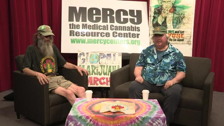 Red curtain background. Mercy cannabis signs in the back. Man in a had and dark shirt seated to the left. Coffee table with tie-dye tablecloth in the center. Man in a blue button up and hat seated to the right