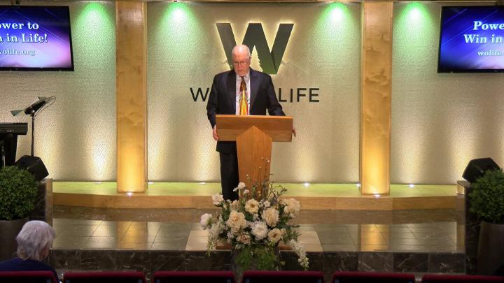 Man in a suit holding a bible next to a pulpit in the middle of a platform. Lit up white walls in the background with a large W and Word of Life in the center.