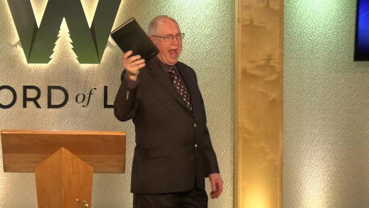 Man in a suit holding a bible next to a pulpit in the middle of a platform. Lit up white walls in the background with a large W and Word of Life in the center.