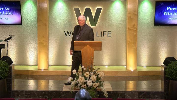 Man in a suit holding a bible next to a pulpit in the middle of a platform. Lit up white walls in the background with a large W and Word of Life in the center.