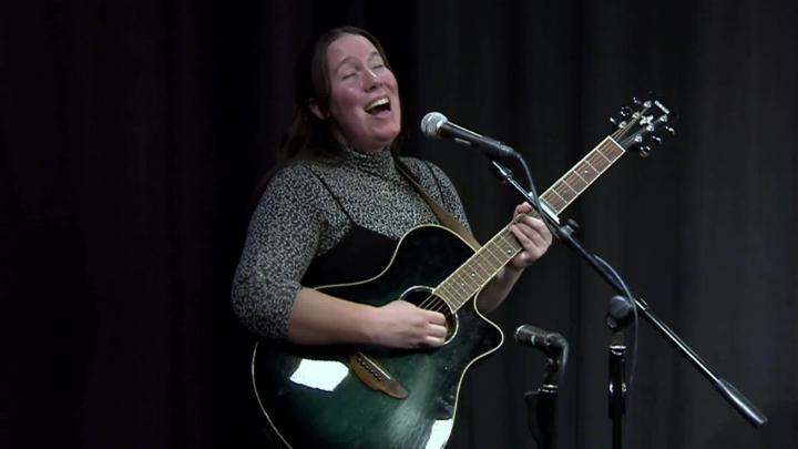 Woman standing in front of a black curtain holding a guitar and singing 