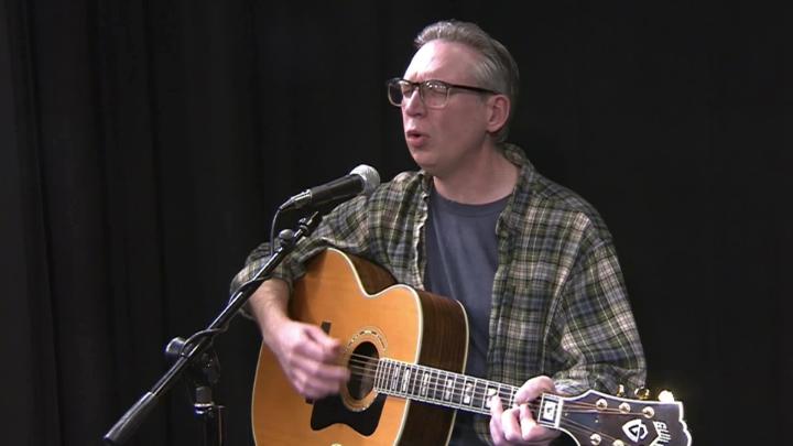 black background, man in a plaid shirt and glasses playing a guitar in front of a mic