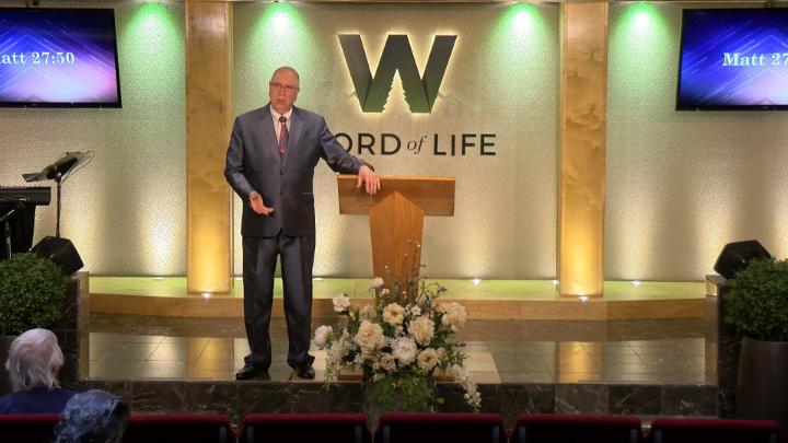 Man in a suit holding a bible next to a pulpit in the middle of a platform. Lit up white walls in the background with a large W and Word of Life in the center.