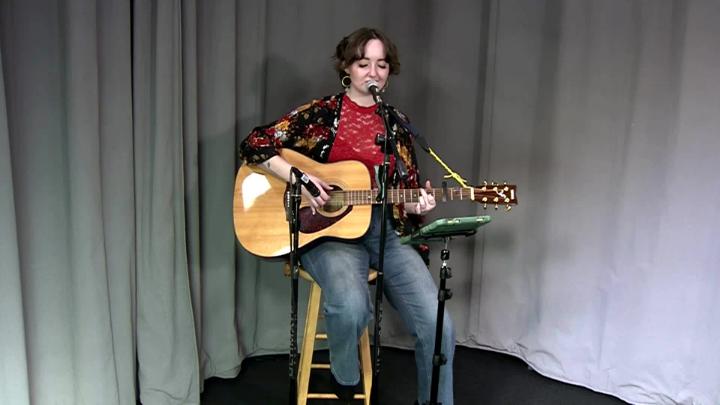 Grey curtain background. Woman with short hair wearing jeans and a red shirt seated behind a mic playing guitar.