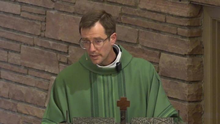 Priest in green standing in front of a brick wall.