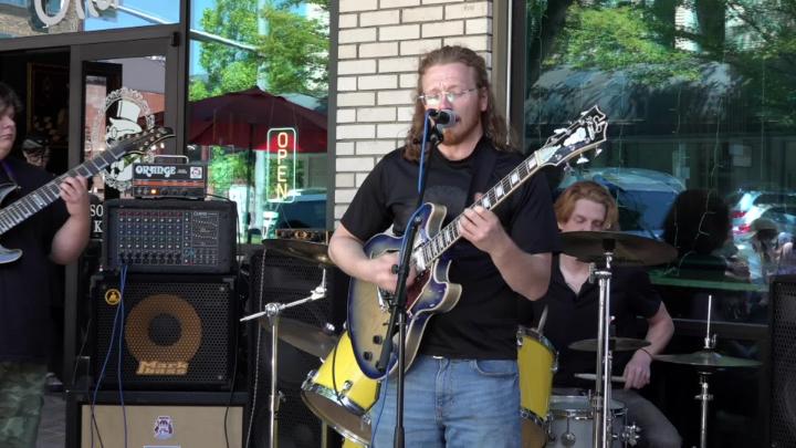 Man in black playing a guitar and singing in front of a brick building