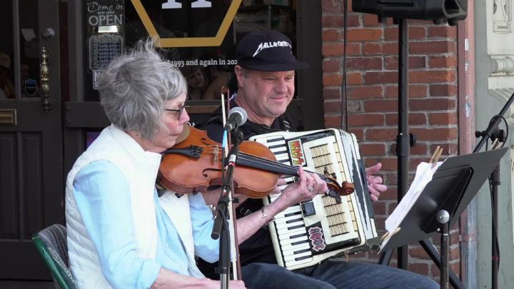 Woman in blue to the left playing a violin. Man to the right in a black hat seated playing an accordian outside