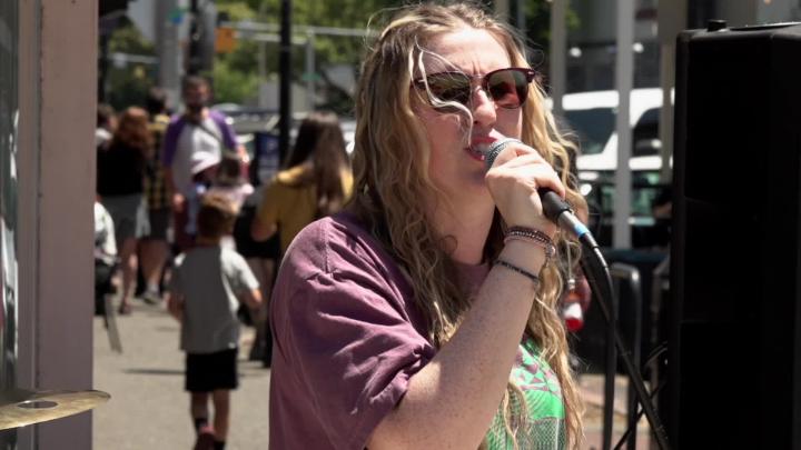 Woman wearing a maroon shirt and sunglasses outside singing into a mic