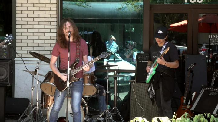 Outside with a band playing. Man in reddish tee standing to the left in front of a mic playing a guitar and singing. Man in dark tee and hat standing off to the right side playing guitar.
