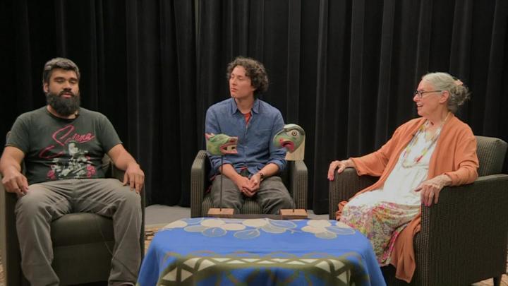 Black curtain background. Man in a tee seated to the left. Man in a blue shirt seated in the center. Woman in a white dress and orange sweater seated to the right.