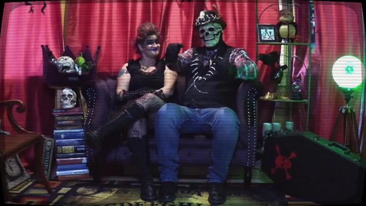 Red curtain background with scifi oddities in the background. A woman is seated on a couch next to a person in a skull mask.