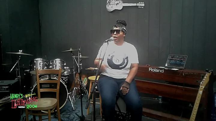Dark wall background. Drums and other instruments behind a woman in a white shirt and jeans seated behind a mic singing.