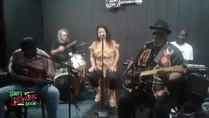 Grey wall in the background. Various musicians seated on stage playing instruments.