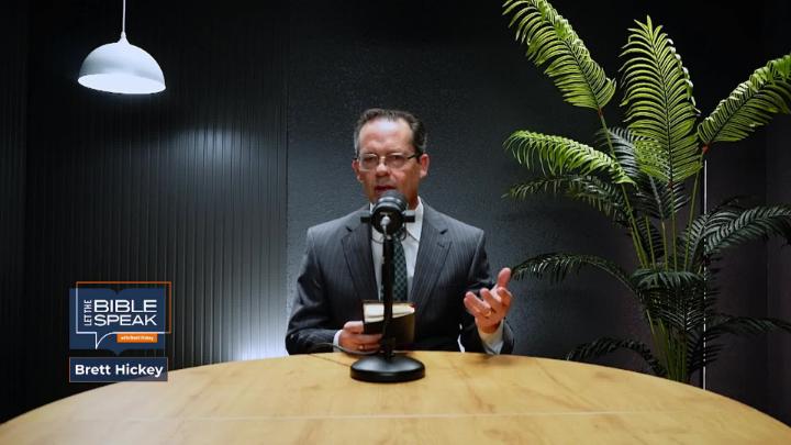 Man in a dark suit sitting at a light colored table in front of a podcast mic. A light is in the top left corner and a house plant to the right. Let the Bible Speak logo bottom left corner.