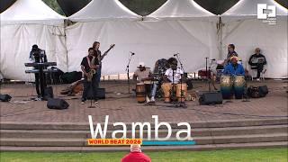 Group of musicians on a brick floored stage with white tents in the background. The word "Wamba" at the bottom.