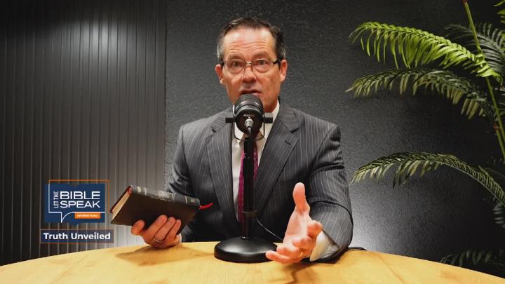 Man in a grey suit seated at a light colored table behind a podcast mic. He is holding a bible. Dark colored walls in the background and a house plant to the right. "Let  The Bible Speak" logo bottom left