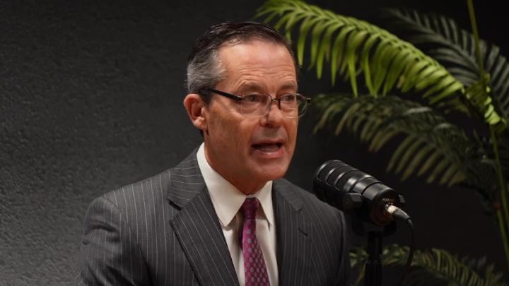 A white man in a grey suit, white shirt, and red tie speaks into a microphone in front of a black background. On the right a green fern sits behind him.