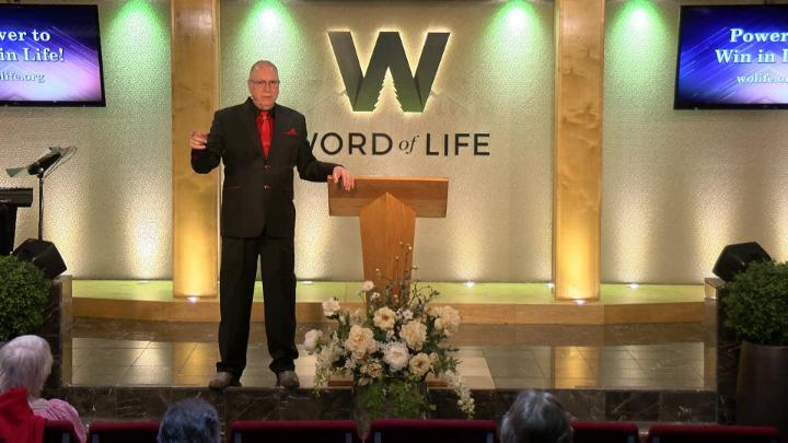 Man in a suit holding a bible next to a pulpit in the middle of a platform. Lit up white walls in the background with a large W and Word of Life in the center.
