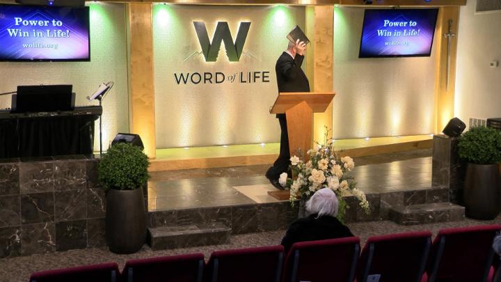 Man in a suit holding a bible next to a pulpit in the middle of a platform. Lit up white walls in the background with a large W and Word of Life in the center.