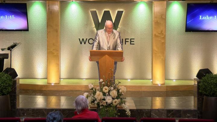 Man in a suit holding a bible next to a pulpit in the middle of a platform. Lit up white walls in the background with a large W and Word of Life in the center.