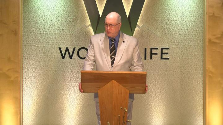 Man in a suit holding a bible next to a pulpit in the middle of a platform. Lit up white walls in the background with a large W and Word of Life in the center.