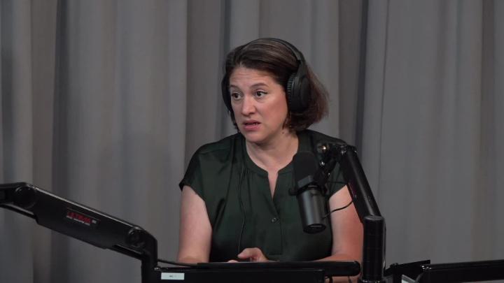 Grey background with a woman dressed in a dark top seated in front of a podcast mic